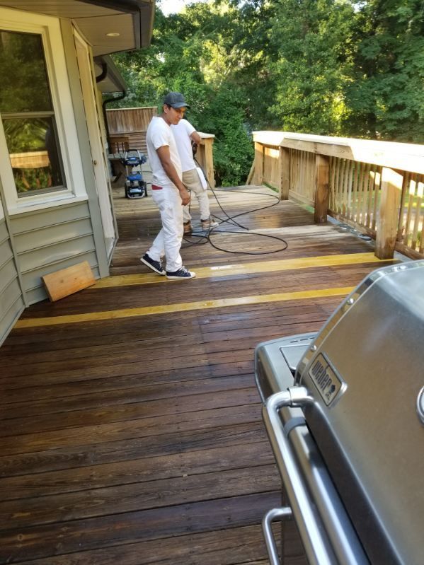 Two people working on a wooden deck; one uses a machine. A grill sits in the foreground.