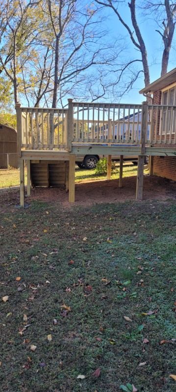 A wooden deck with a railing stands in a yard with sparse grass under a clear blue sky.