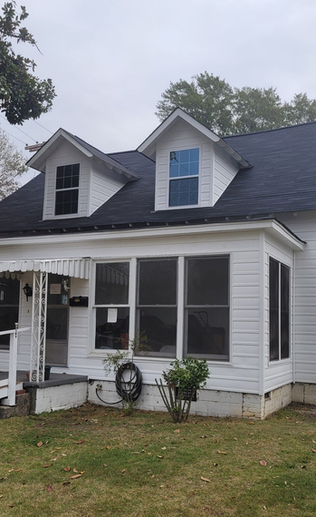 White house with black roof, two dormers, and a porch. Overcast sky.
