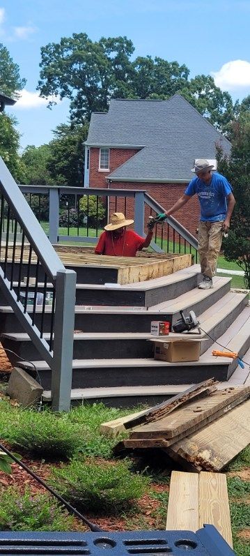 Two workers building a deck. One in a red shirt and straw hat, another in a blue shirt. Steps are in progress.