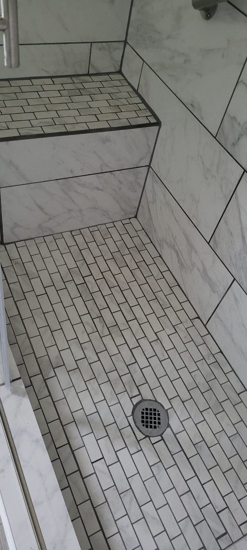 Tiled shower stall with a built-in bench. The floor has a drain in the center. White with black grout.
