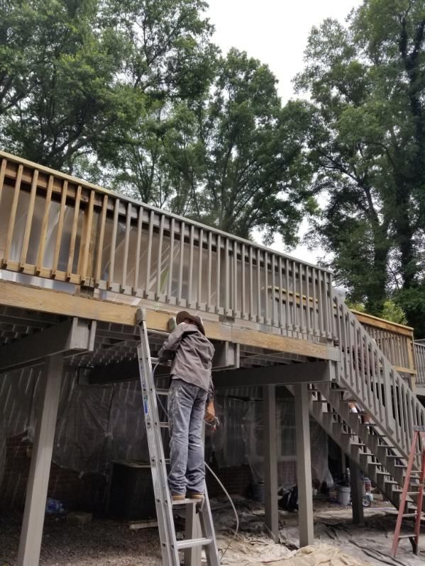 Person on ladder works on deck; gray railings, wood construction, trees in background.