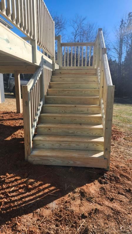 Wooden outdoor staircase leading up to a deck, set on reddish soil, against a blue sky.