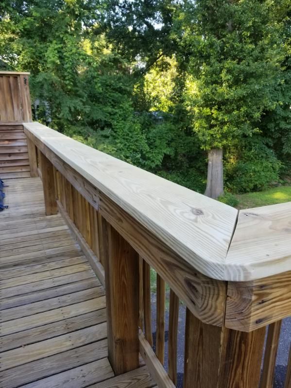 Wooden deck railing with light wood top rail and vertical supports, set against lush green trees.
