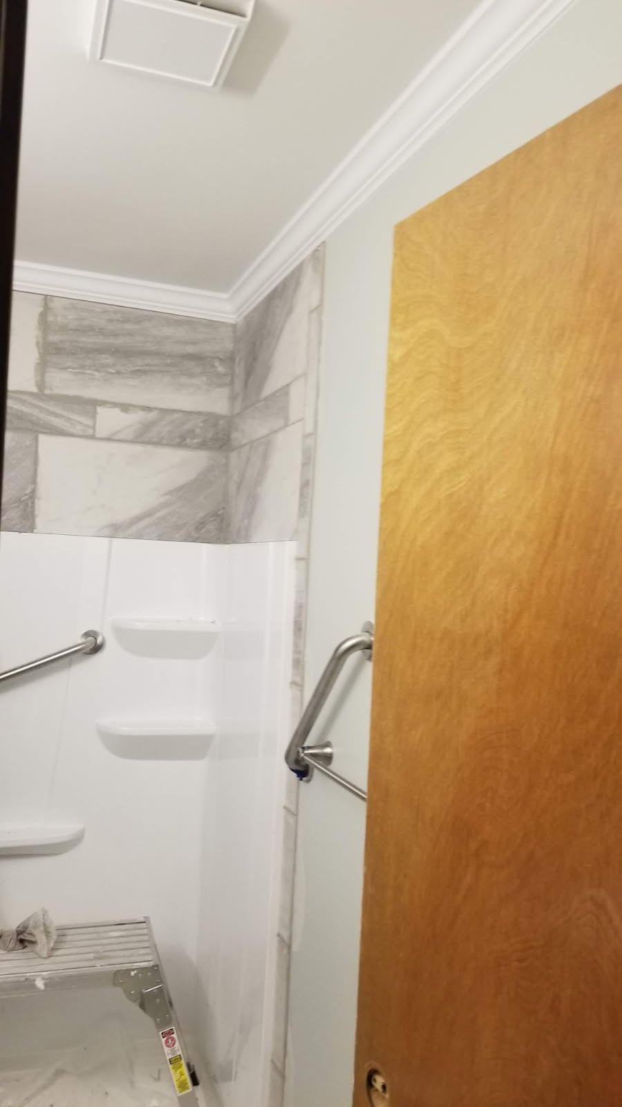Bathroom with white shower stall, marble-look tiled wall, grab bars, and a wooden door.