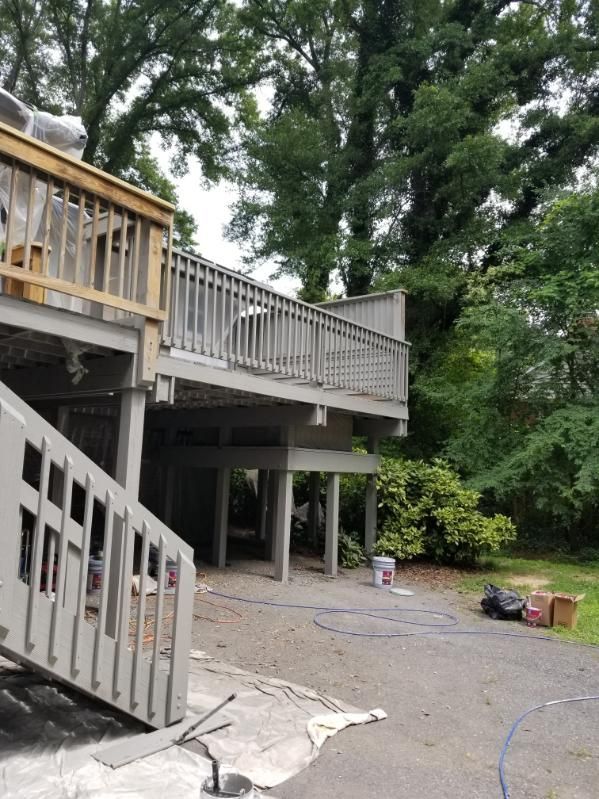 Elevated wooden deck painted gray, with stairs leading down. Surrounded by trees and a paved area.