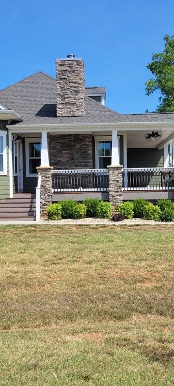 House with a stone chimney, porch with white columns, and green bushes, sunny day.