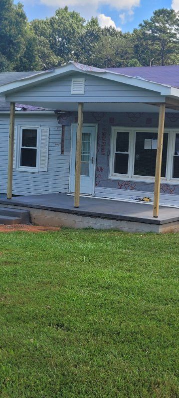 A light blue house with a covered porch under construction, green lawn, trees, and cloudy blue sky.