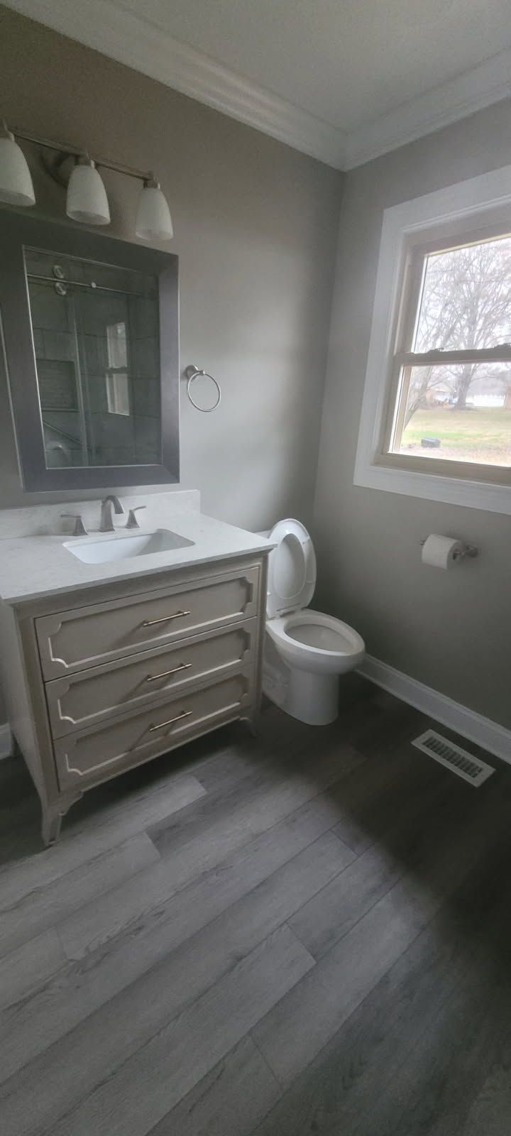Bathroom with gray walls, light wood floor, toilet, vanity, and window.
