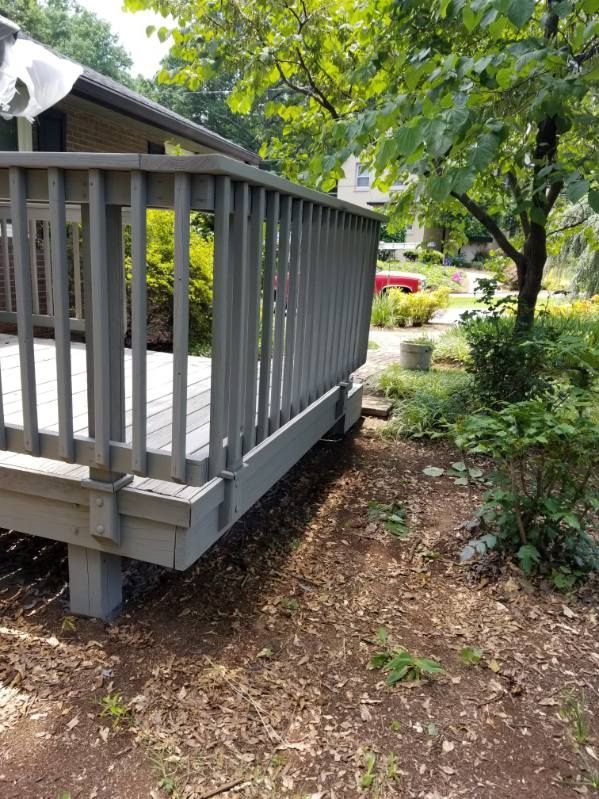 Gray deck with railing, supported by posts, in a yard with trees and mulch.