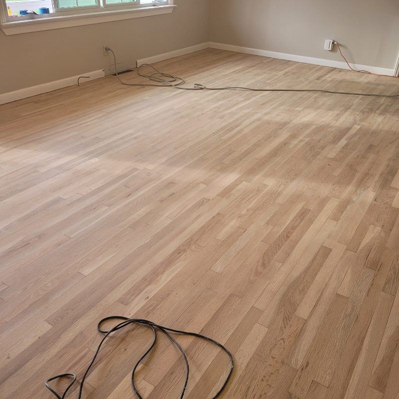 Light-colored wood floor in a mostly empty room with beige walls. Cords lie on the floor.