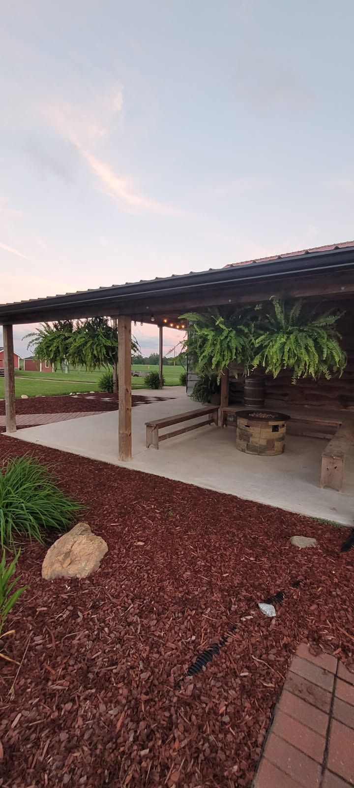 Outdoor wooden structure with bench and fire pit, set on a concrete pad with surrounding reddish-brown mulch.