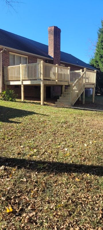 A two-level wooden deck attached to a brick house with a chimney and stairs, in a grassy backyard.