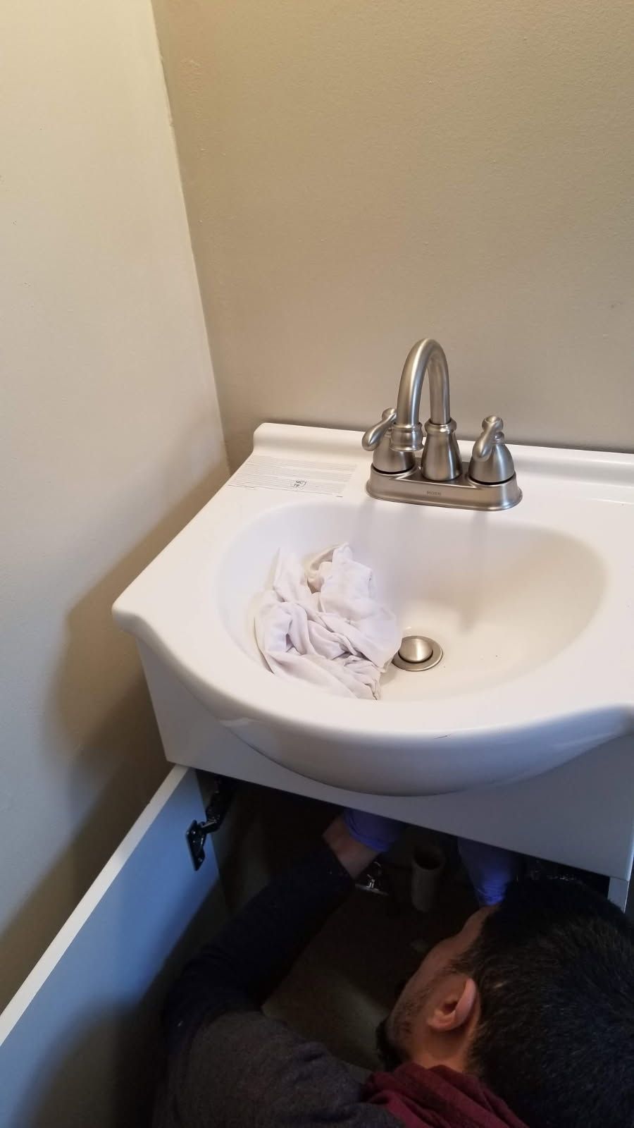Person working under a white bathroom sink, with a partially open cabinet. Beige walls.