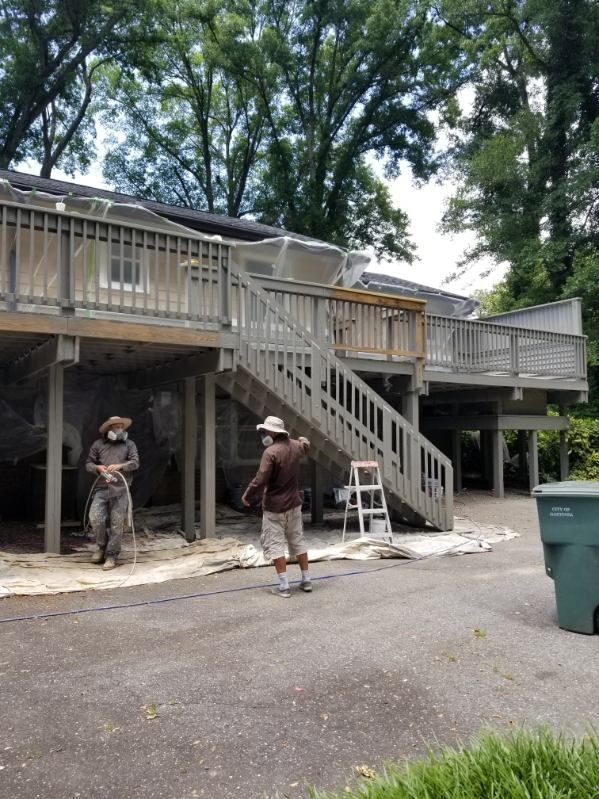 Two workers near a two-story wooden deck, one holding a tool, another wearing a hat.