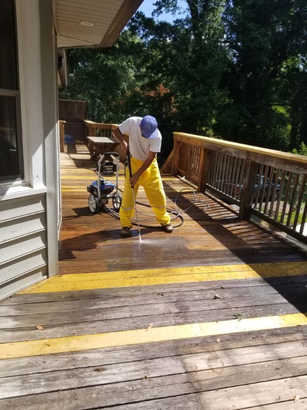 Person in yellow protective gear pressure washing a wooden deck on a sunny day.