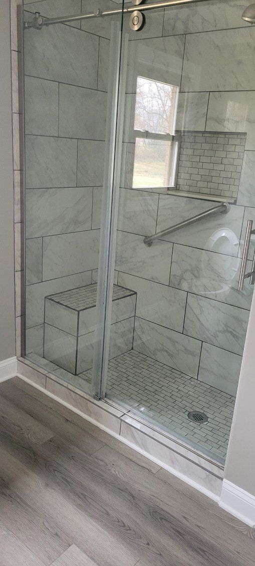 Glass shower with tiled walls and a bench, set in a bathroom with gray flooring.