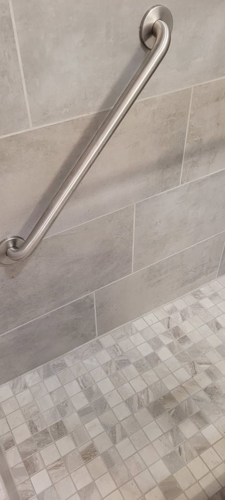 A stainless steel grab bar mounted on a tiled wall above a mosaic tiled floor.