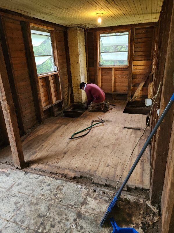 A person kneels in a room with exposed wooden walls and floor, two windows, and a blue broom.