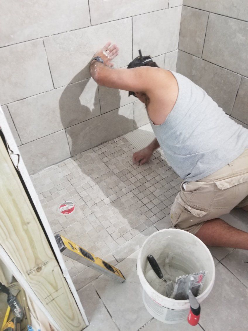 A person tiling a shower floor, using tools like a level and bucket, inside a partially tiled shower stall.