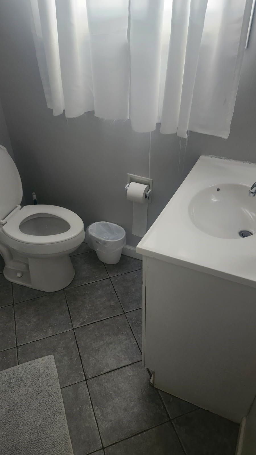 Bathroom with a toilet, sink, wastebasket, and window with white curtains. Grey tile floor.