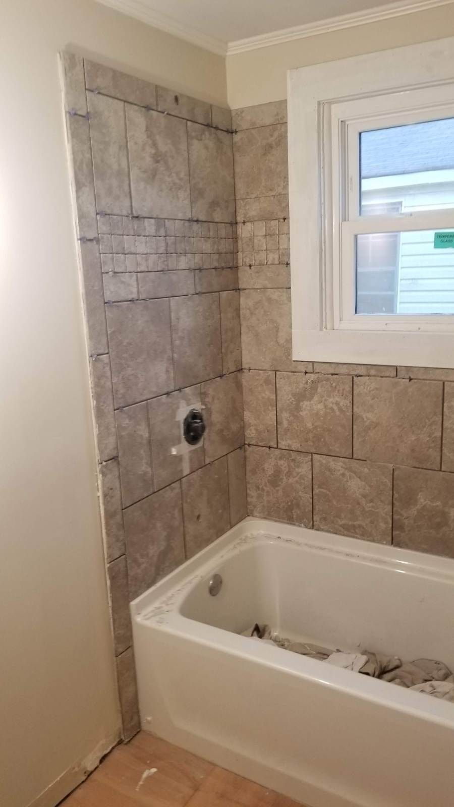 Bathroom shower wall partially tiled with brown stone-like tiles, white bathtub, window.