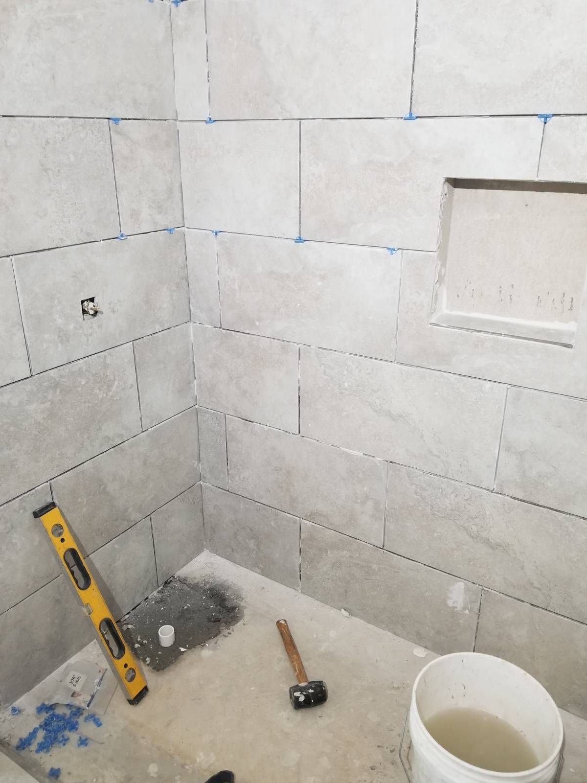 A tiled shower under construction. Gray tiles with blue tape, tools on floor: level, trowel, hammer, bucket.