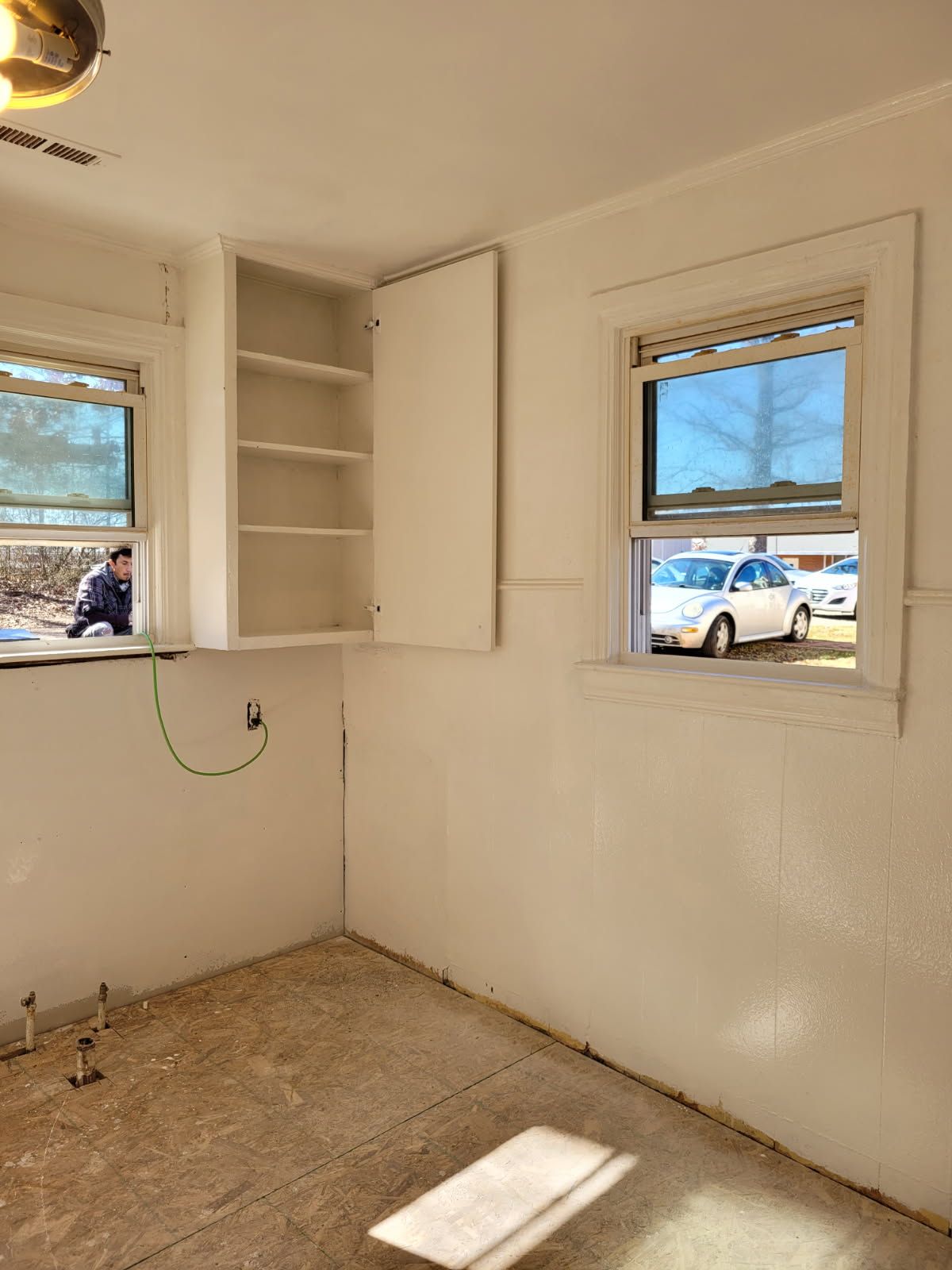 Empty kitchen with white walls, cabinets, windows, and exposed floor. A person is visible outside a window.