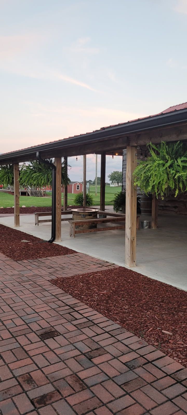 A covered outdoor seating area with tables and brick pathway. Wooden beams, mulch, and greenery are visible.