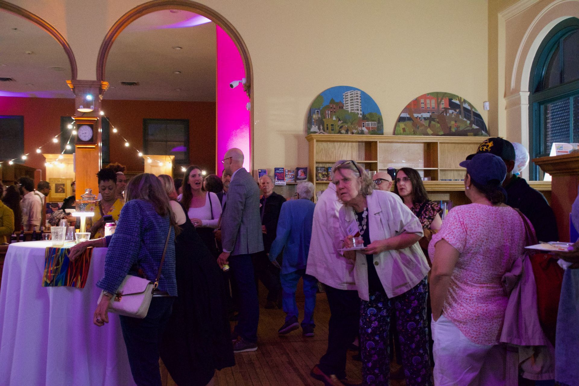 People in formal attire gather for a social event in a well-lit hall with arched doorways, string lights, and wall art.