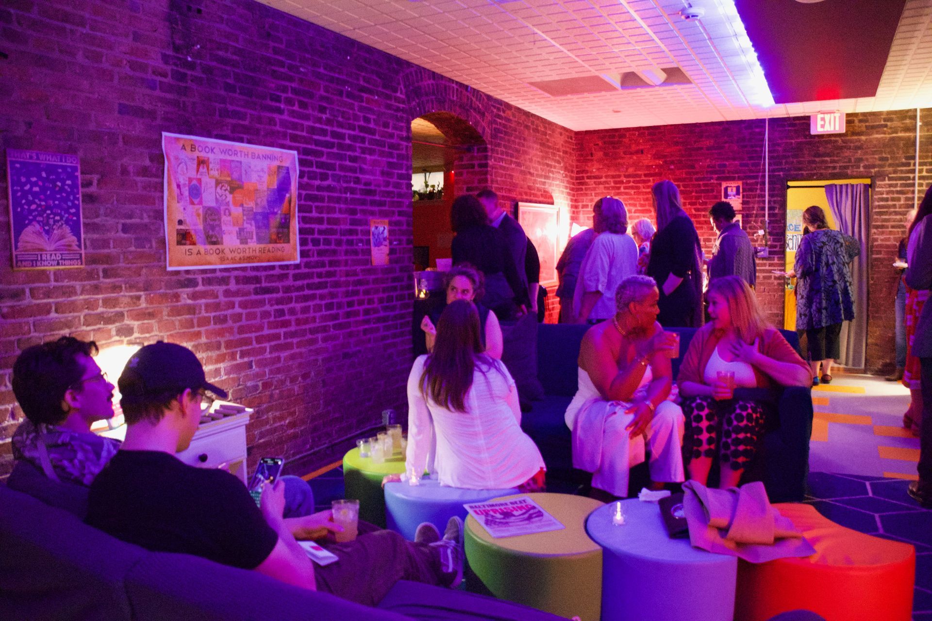 People socialize in a room with brick walls, purple lighting, and brightly colored round stools and chairs.