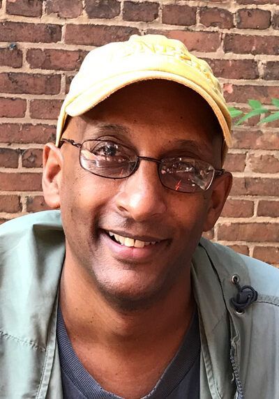 A man wearing glasses and a yellow hat is smiling in front of a brick wall.
