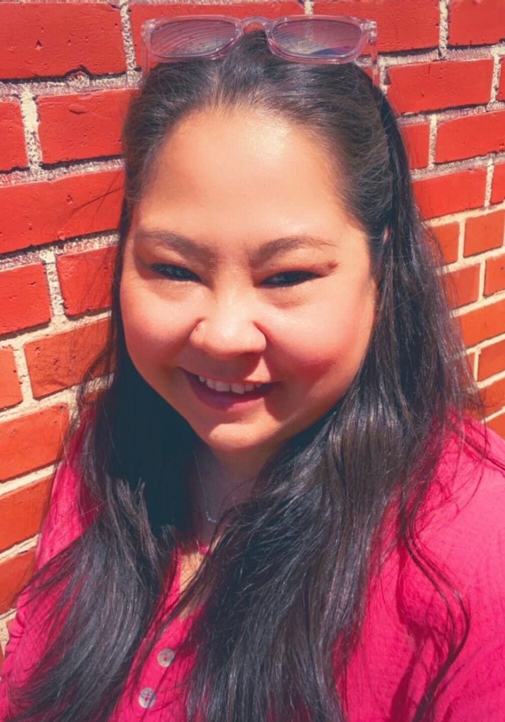 A woman wearing sunglasses is smiling in front of a brick wall.