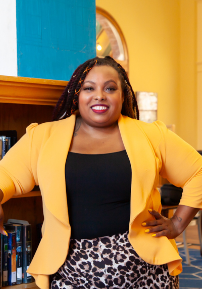 A woman in a yellow jacket and leopard print skirt is standing in front of a bookshelf.