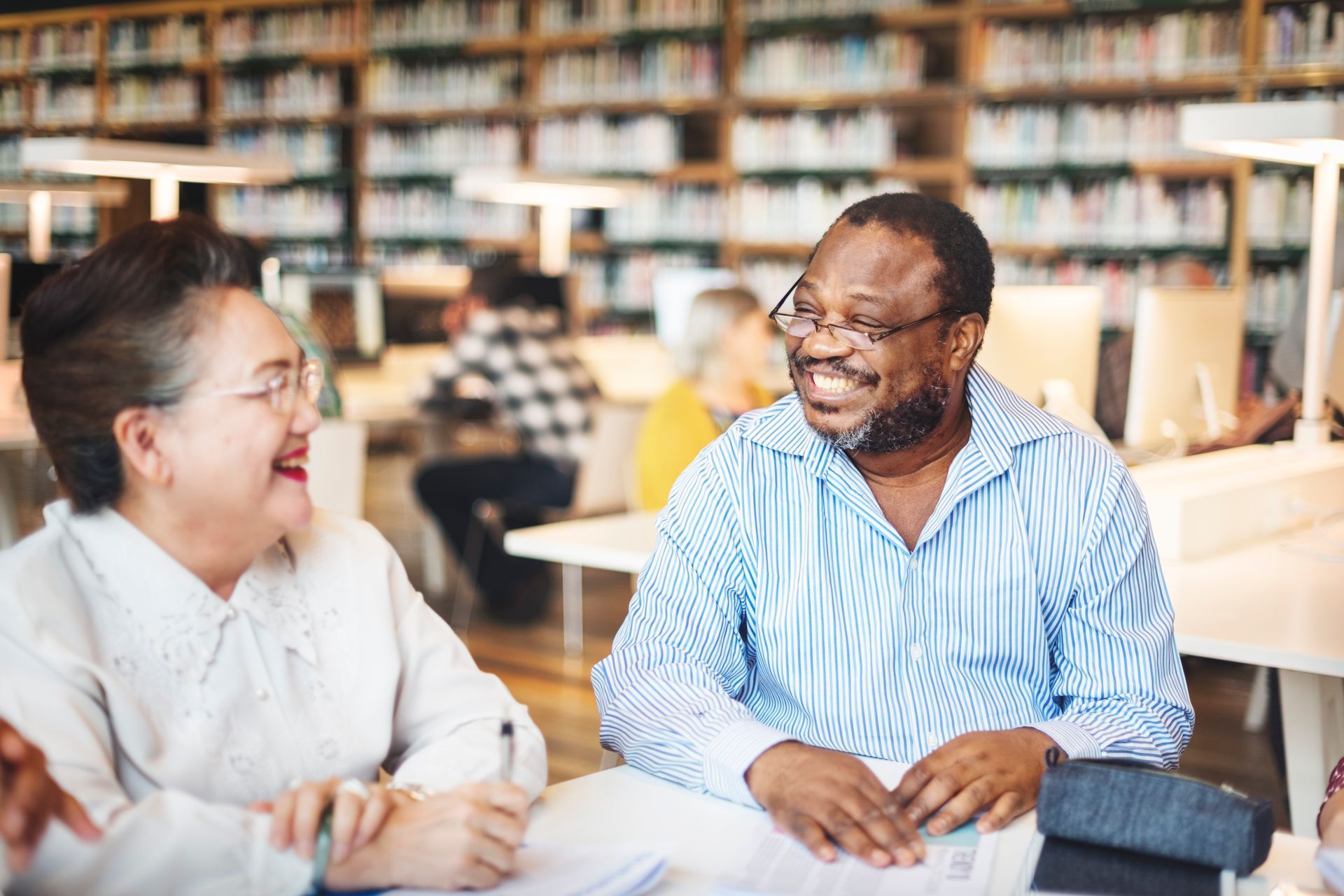 A man and a woman are sitting at a table in a library talking to each other.