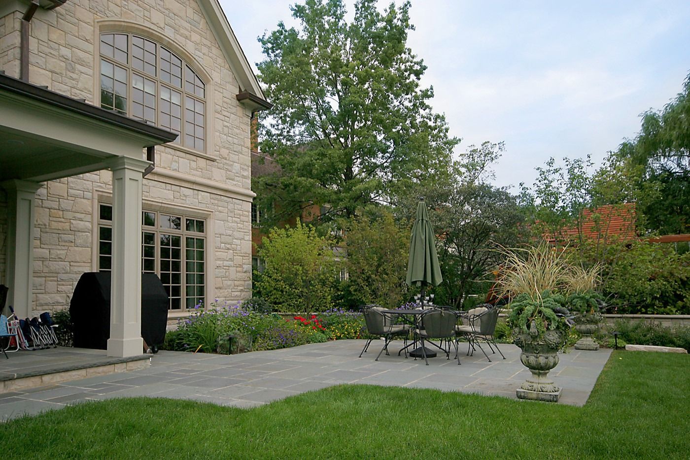 Stone house patio with outdoor table and chairs, potted plants, and a green lawn in a landscaped garden.