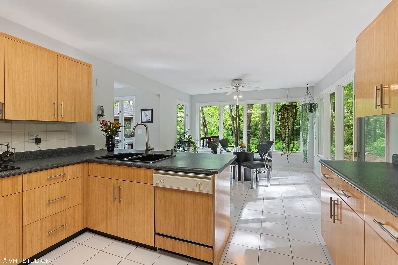 Bright kitchen with wooden cabinets, black countertops, and a view into a sunlit dining area with glass doors.