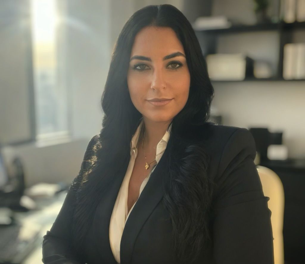 Woman with dark hair, wearing a black blazer and white top, looking at the camera. Sitting in an office.