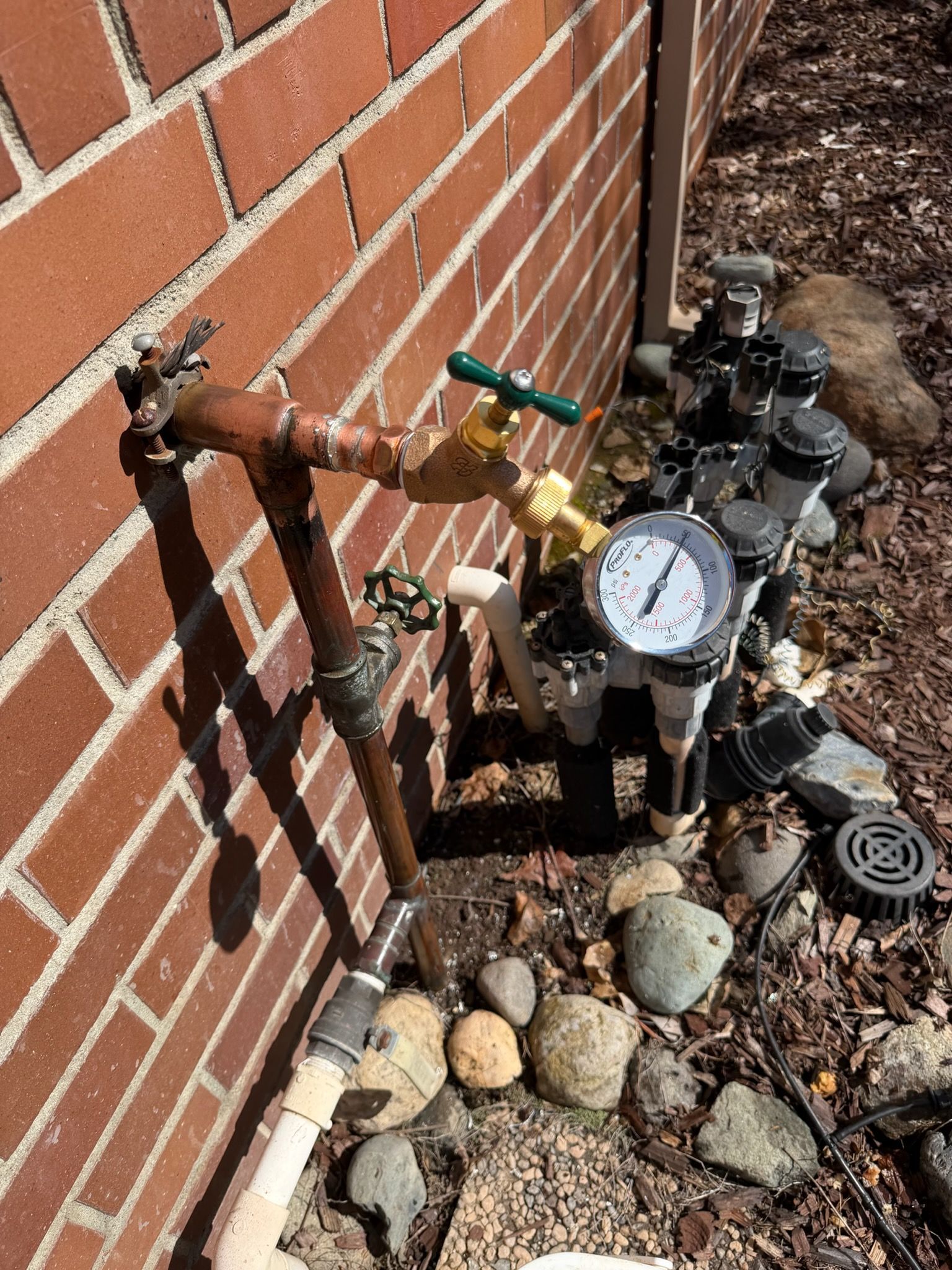 An outdoor irrigation system setup with a pressure gauge and valves mounted against a brick wall on a bed of rocks.