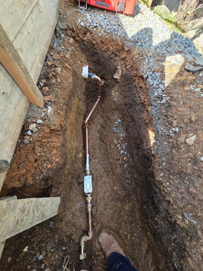 Copper gas line in a trench next to a concrete wall, with a foot visible in the bottom right corner.