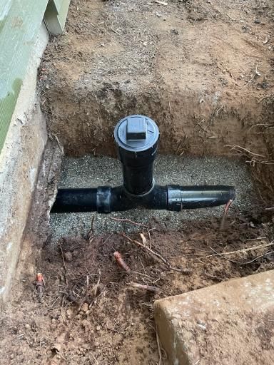 Black plumbing pipes and fitting in a dirt pit, near a building foundation.