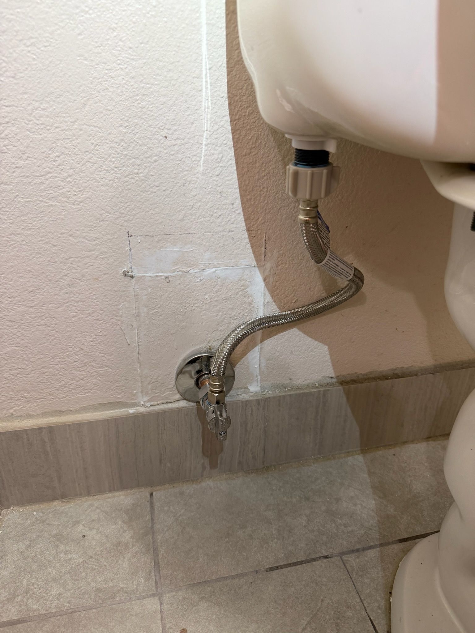 A stainless steel supply hose connects a white toilet tank to a chrome shut-off valve on a tiled bathroom wall.