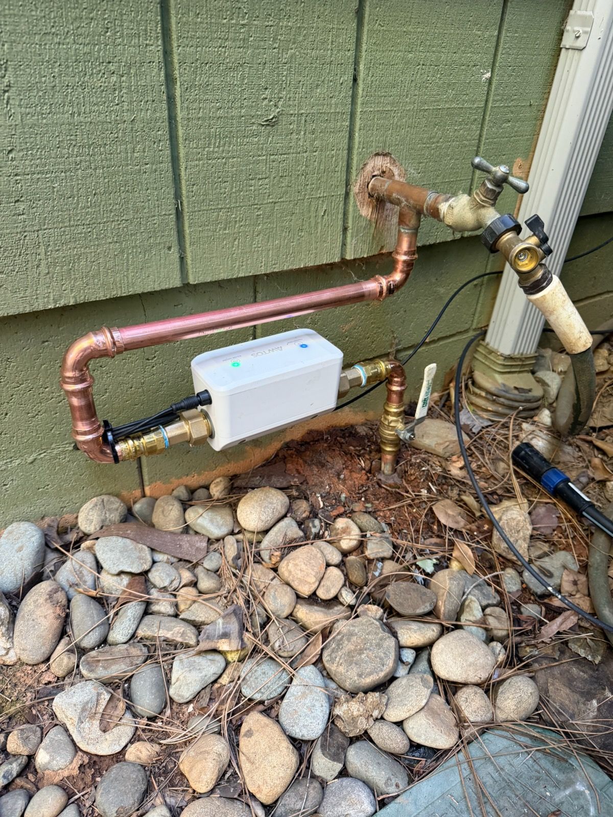 A water pipe installation featuring a white rectangular sensor box mounted on a wall next to a hose spigot over gravel.