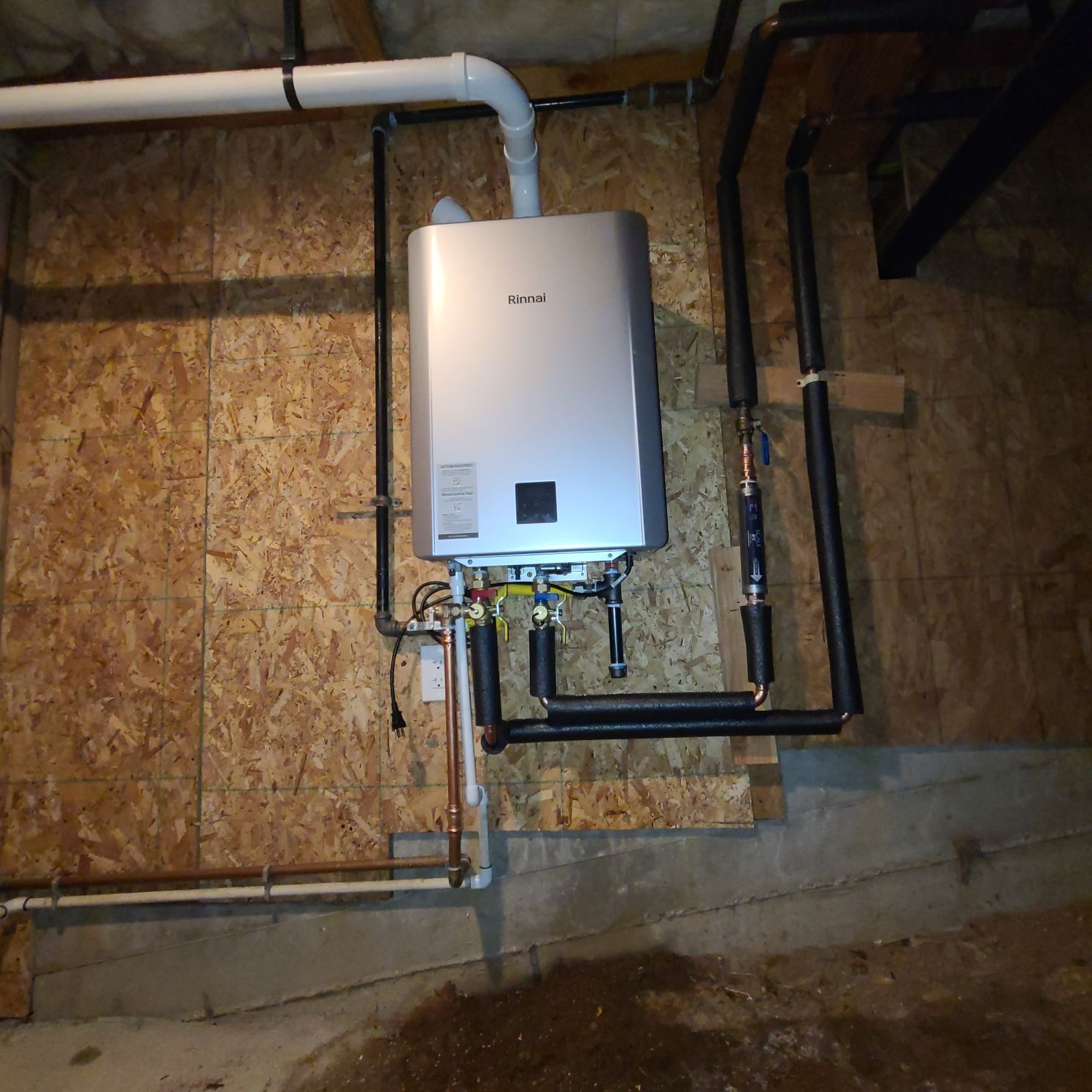 A tankless water heater mounted on a wood-paneled wall in a basement. Black pipes are connected to the silver unit.