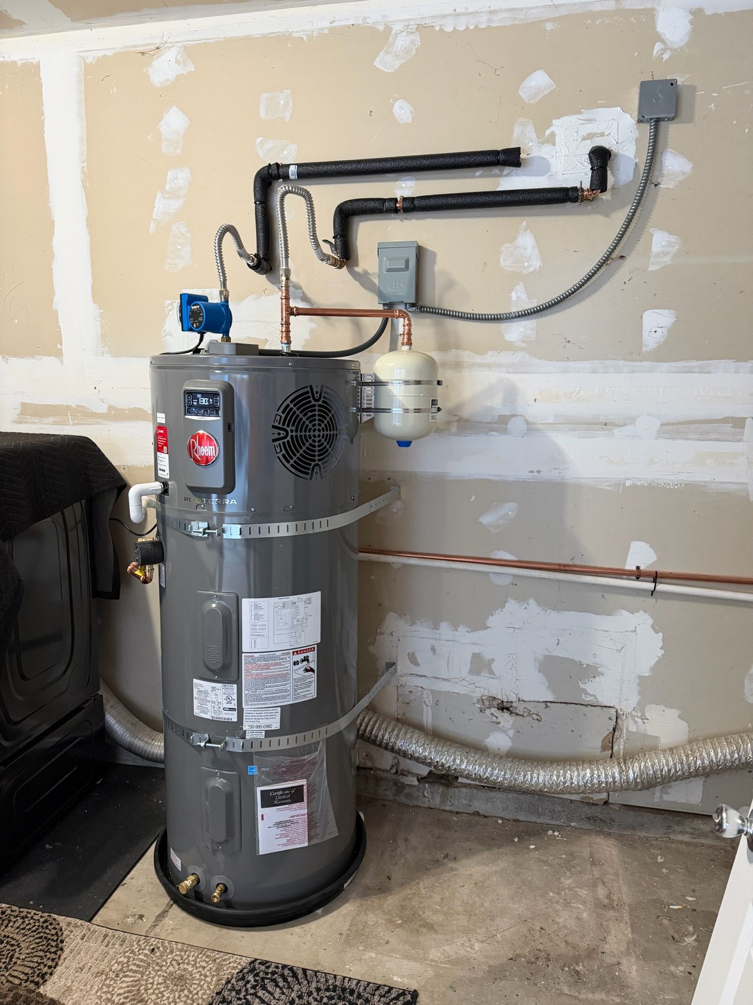 A water heater in a garage with pipes, wiring, and a pressure relief valve.