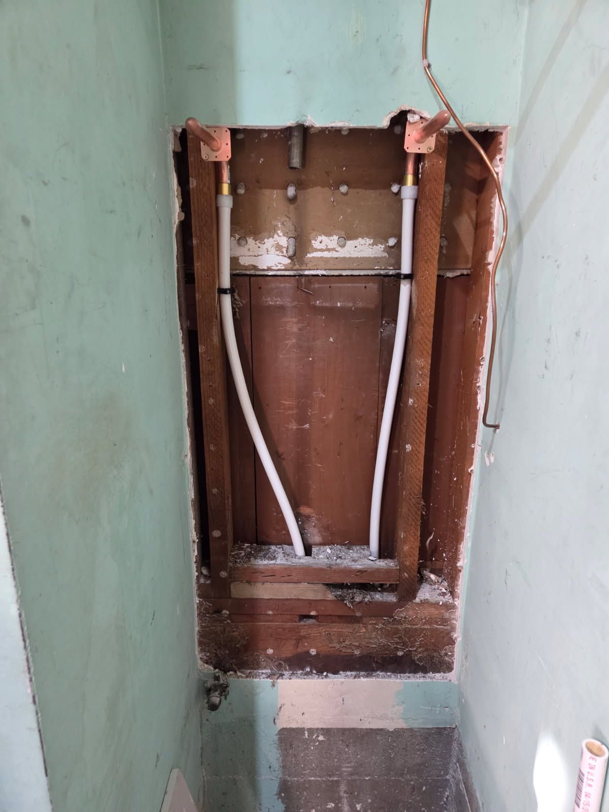 Plumbing pipes in an open wall cavity, white against a brown wooden frame, in a green room.