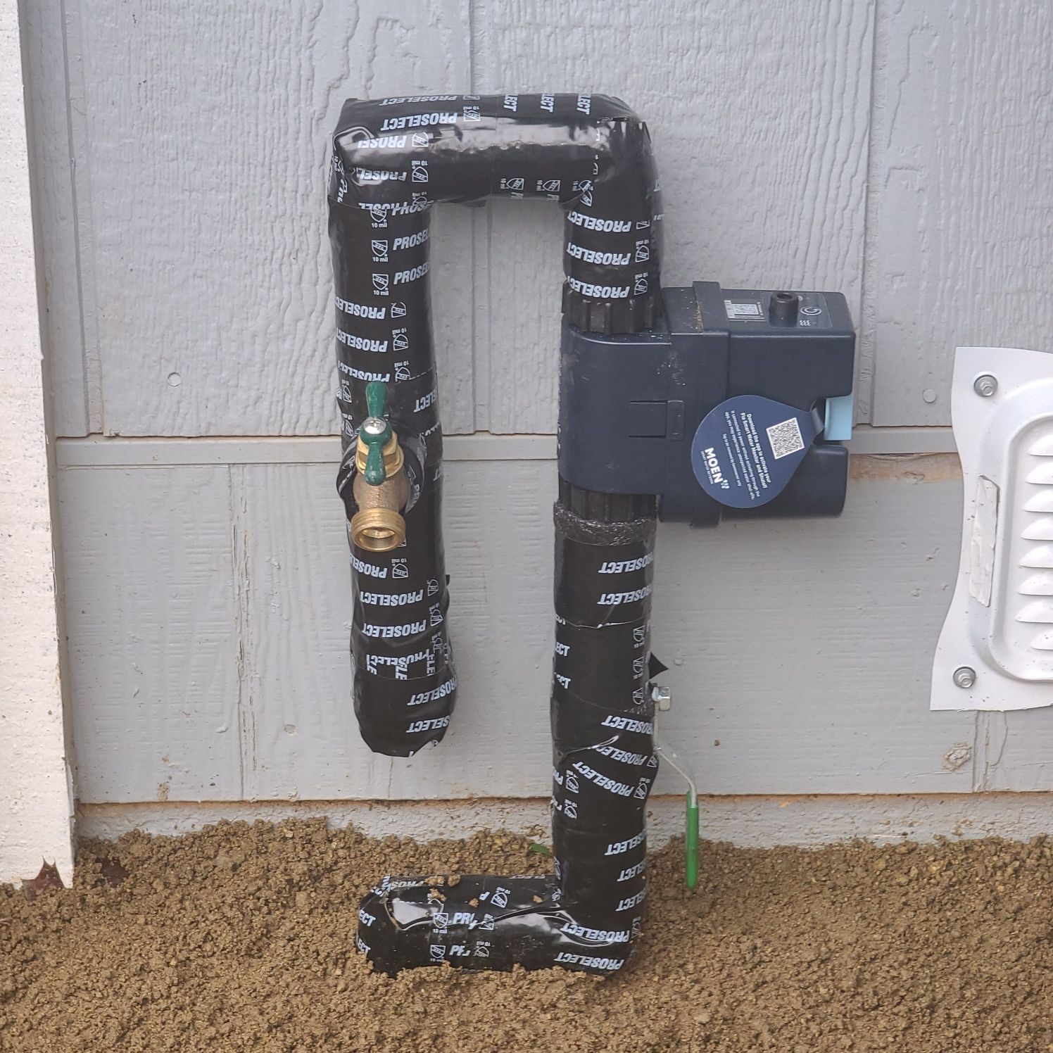 Black plumbing pipes with a pump and valve against a light brown wall, wrapped in black tape, sitting on loose tan ground.