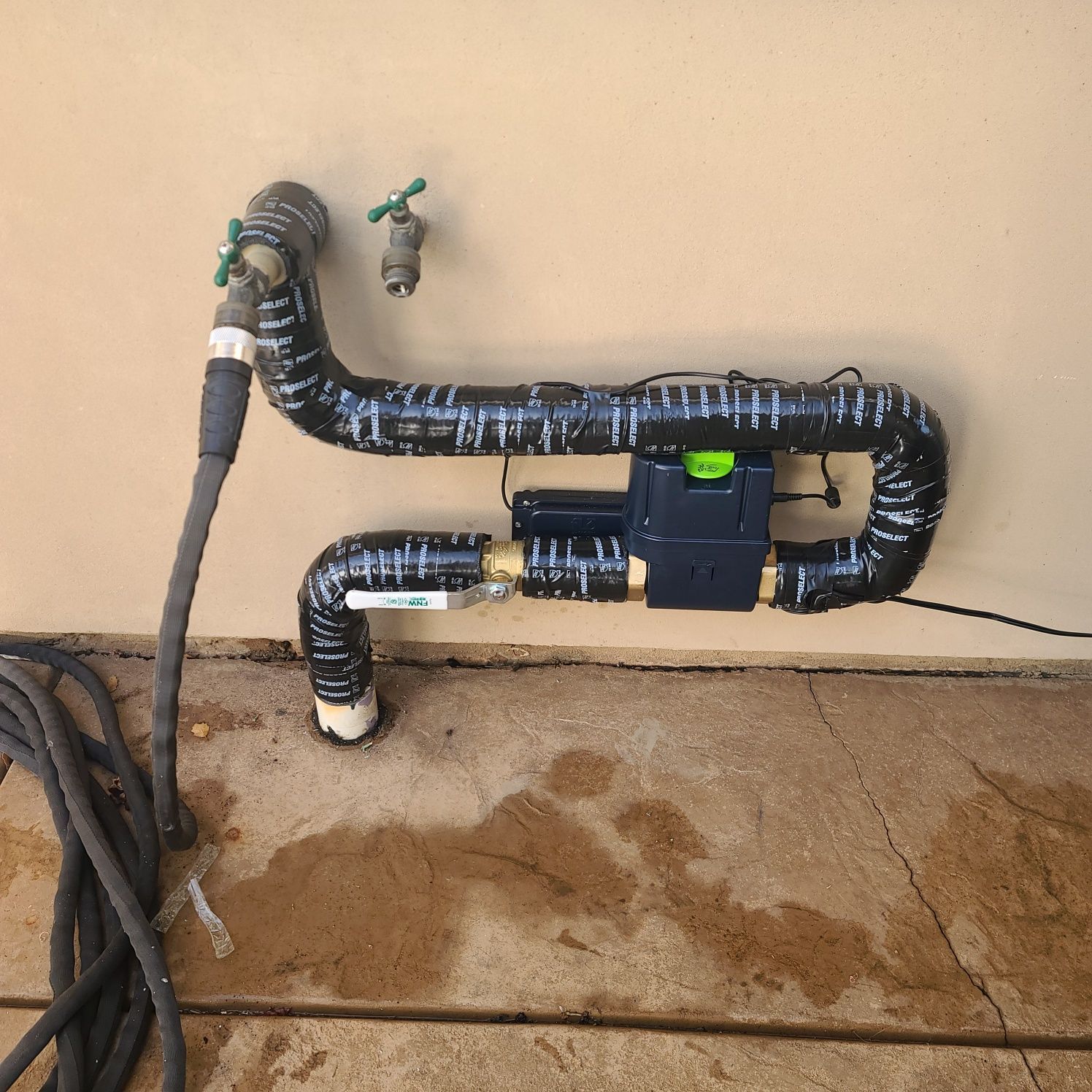 Insulated outdoor pipes and a valve mounted on a beige wall above a wet concrete floor.