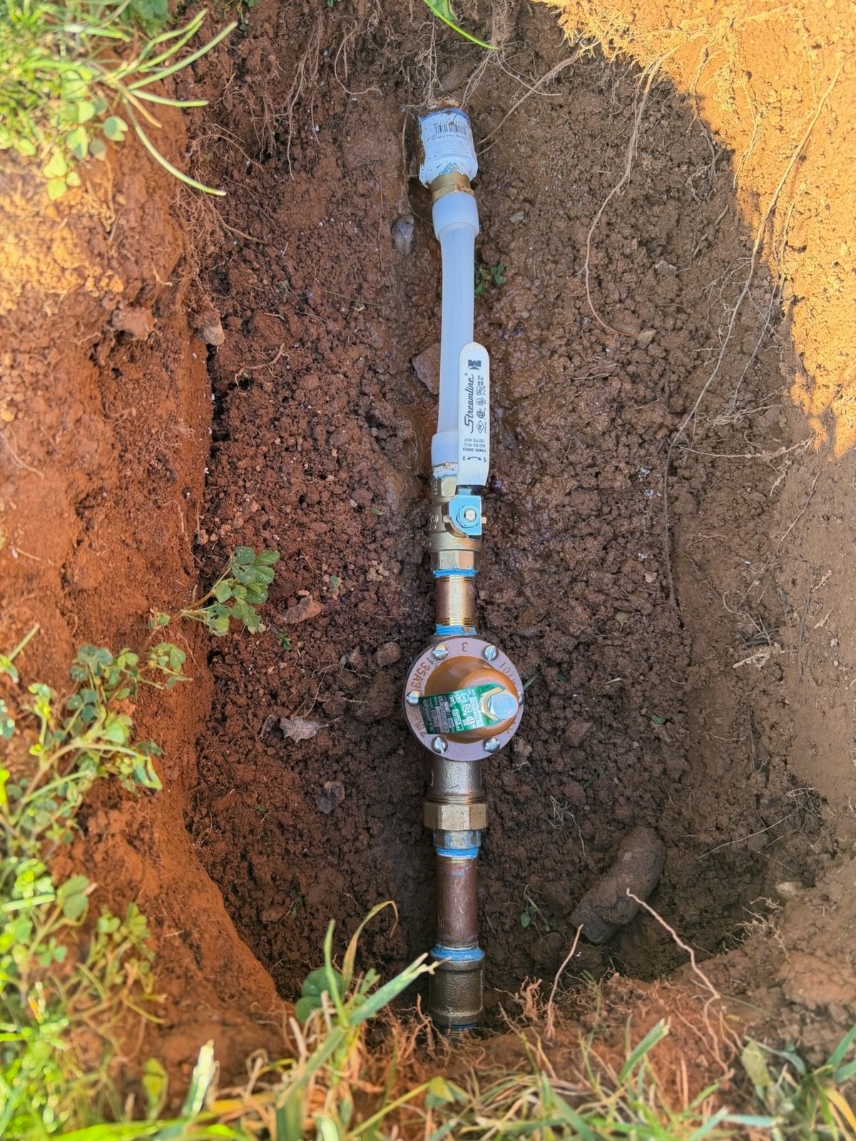 Pipes and water meter installed in a narrow, muddy trench in the ground with surrounding grass and soil.