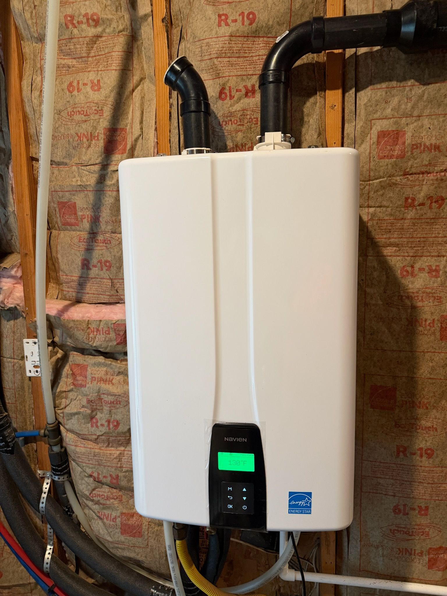 White, wall-mounted tankless water heater with black pipes and a green digital display against insulation.